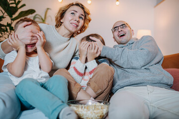 Family watching television together on couch, parents covering children's faces