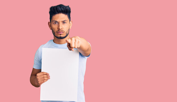 Handsome latin american young man holding cardboard banner with blank space pointing with finger to the camera and to you, confident gesture looking serious