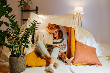 Tender loving mother and daughter sitting in tent with book, touching foreheads