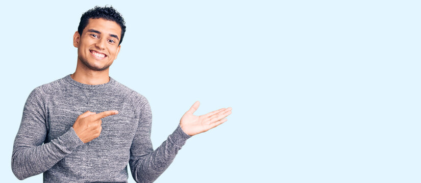 Hispanic handsome young man wearing casual sweater amazed and smiling to the camera while presenting with hand and pointing with finger.