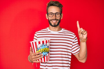 Handsome caucasian man with beard eating popcorn smiling with an idea or question pointing finger with happy face, number one