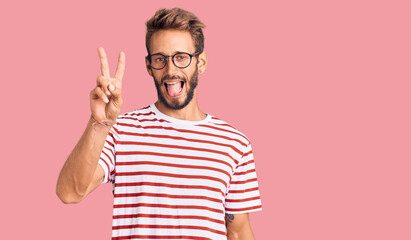 Handsome blond man with beard wearing casual clothes and glasses smiling with happy face winking at the camera doing victory sign with fingers. number two.