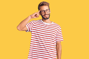 Handsome blond man with beard wearing casual clothes and glasses smiling pointing to head with one finger, great idea or thought, good memory