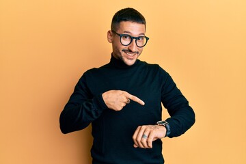 Handsome man with tattoos wearing turtleneck sweater and glasses in hurry pointing to watch time,...
