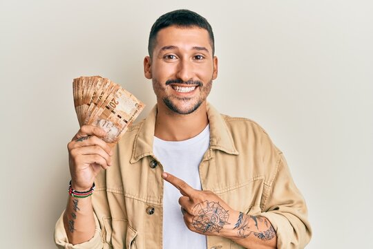 Handsome man with tattoos holding south african 20 rand banknotes smiling happy pointing with hand and finger
