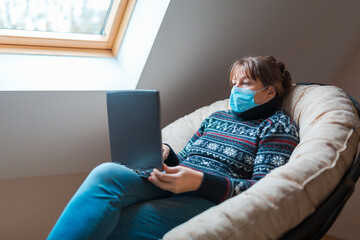 Obraz premium The woman is in self isolation in the house. Remote work on a laptop computer sitting in a chair.