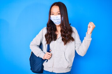 Young beautiful chinese girl holding student backpack wearing medical mask screaming proud, celebrating victory and success very excited with raised arms