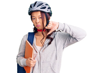 Young beautiful chinese girl wearing student backpack and bike helmet holding book with angry face, negative sign showing dislike with thumbs down, rejection concept