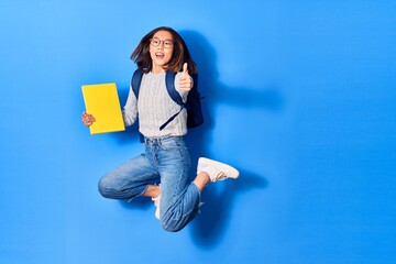 Young beautiful chinese student girl wearing glasses and backpack smiling happy. Jumping with smile on face holding book doing ok sign over isolated blue background