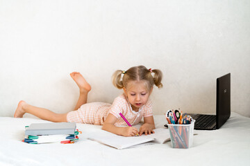 Little girl performs a task in a notebook on the bed, remote learning, home learning concept