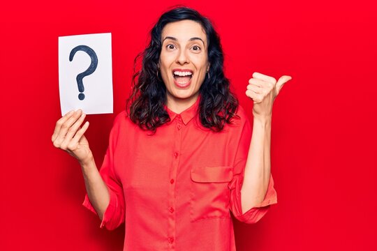 Young beautiful hispanic woman holding question mark pointing thumb up to the side smiling happy with open mouth