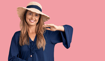 Middle age hispanic woman wearing summer hat gesturing with hands showing big and large size sign, measure symbol. smiling looking at the camera. measuring concept.