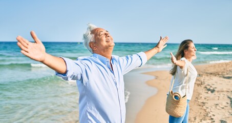 Middle age hispanic couple breathing with arms raised at the beach.