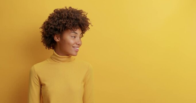 Pretty curly haired woman smiles broadly and turns head away looks on blank space over yellow wall reads pleasant announcement sees awesome advertisement rejoices sale dressed in casual wear