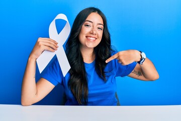 Beautiful brunette young woman holding white ribbon pointing finger to one self smiling happy and proud