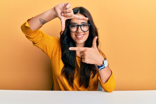 Beautiful brunette young woman wearing glasses and casual clothes sitting on the table smiling making frame with hands and fingers with happy face. creativity and photography concept.