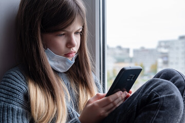 Sad little / young girl in a protective mask sits by the window and holds a phone in her hands