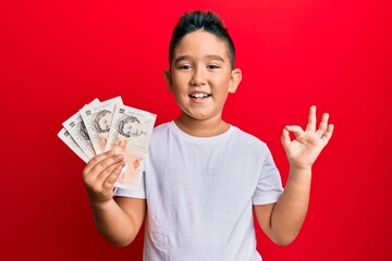 Little boy hispanic kid holding 10 united kingdom pounds banknotes doing ok sign with fingers, smiling friendly gesturing excellent symbol