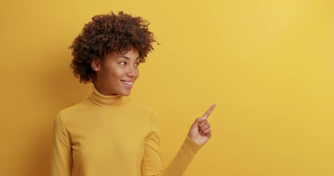 Cheerful positive young Afro American woman points at blank space at empty space shows advertisement with glad expression nods head and suggests to try product poses against yellow background