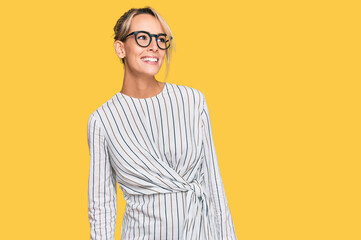 Beautiful blonde woman wearing business shirt and glasses looking away to side with smile on face,...