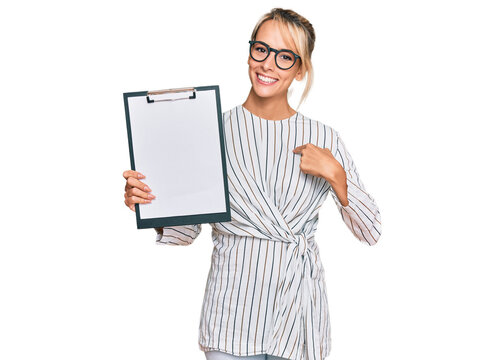 Beautiful blonde business woman holding clipboard with blank space pointing finger to one self smiling happy and proud