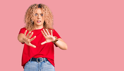 Young blonde woman with curly hair wearing casual red tshirt afraid and terrified with fear expression stop gesture with hands, shouting in shock. panic concept.