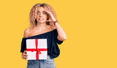 Young blonde woman with curly hair holding gift stressed and frustrated with hand on head, surprised and angry face