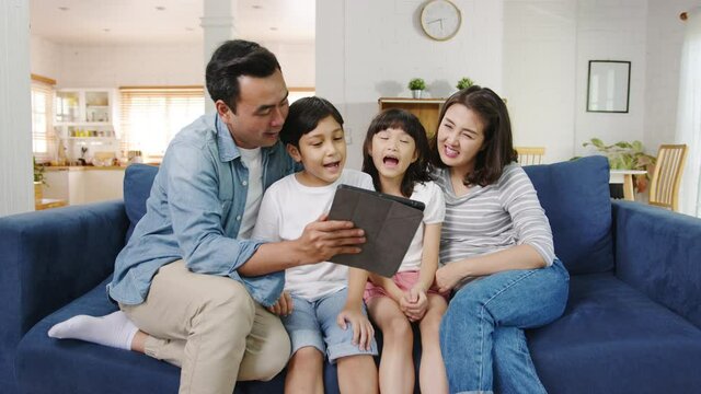 Happy cheerful Asian family dad, mom and kids having fun and using digital tablet video call on sofa at house. Self-isolation, stay at home, social distancing, quarantine for coronavirus prevention.