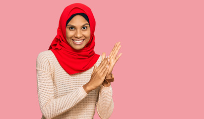 Young african american woman wearing traditional islamic hijab scarf clapping and applauding happy...