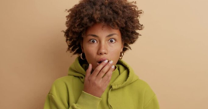 Human reaction concept. Surprised dark skinned woman with curly hair stares shocked at camera keeps mouth widely opened wears casual hoodie isolated over brown background. Embarrassed teenage girl