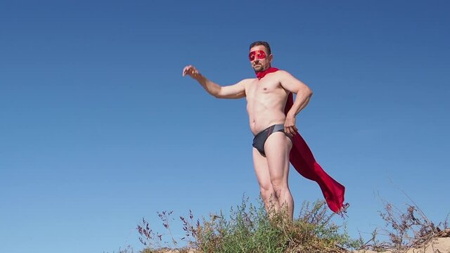 Middle-aged man standing and dressing like superheroes on the beach. Male super hero with red cloak rasing hand on the beach, slow motion.