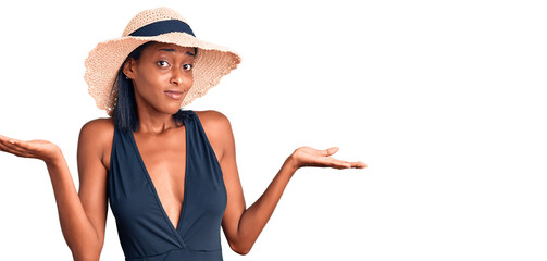 Young african american woman wearing swimsuit and summer hat clueless and confused expression with...