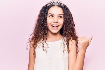 Beautiful kid girl with curly hair wearing princess tiara pointing thumb up to the side smiling happy with open mouth © Krakenimages.com