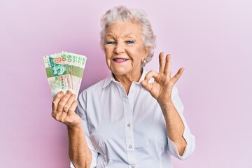 Senior grey-haired woman holding hong kong 50 dollars banknotes doing ok sign with fingers, smiling friendly gesturing excellent symbol