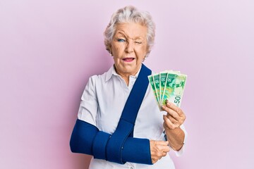 Senior grey-haired woman wearing arm on sling holding israel shekels banknotes winking looking at...