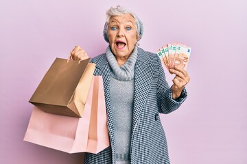 Senior grey-haired woman holding shopping bags and euros banknotes celebrating crazy and amazed for...