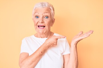 Senior beautiful woman with blue eyes and grey hair wearing classic white tshirt over yellow background amazed and smiling to the camera while presenting with hand and pointing with finger.