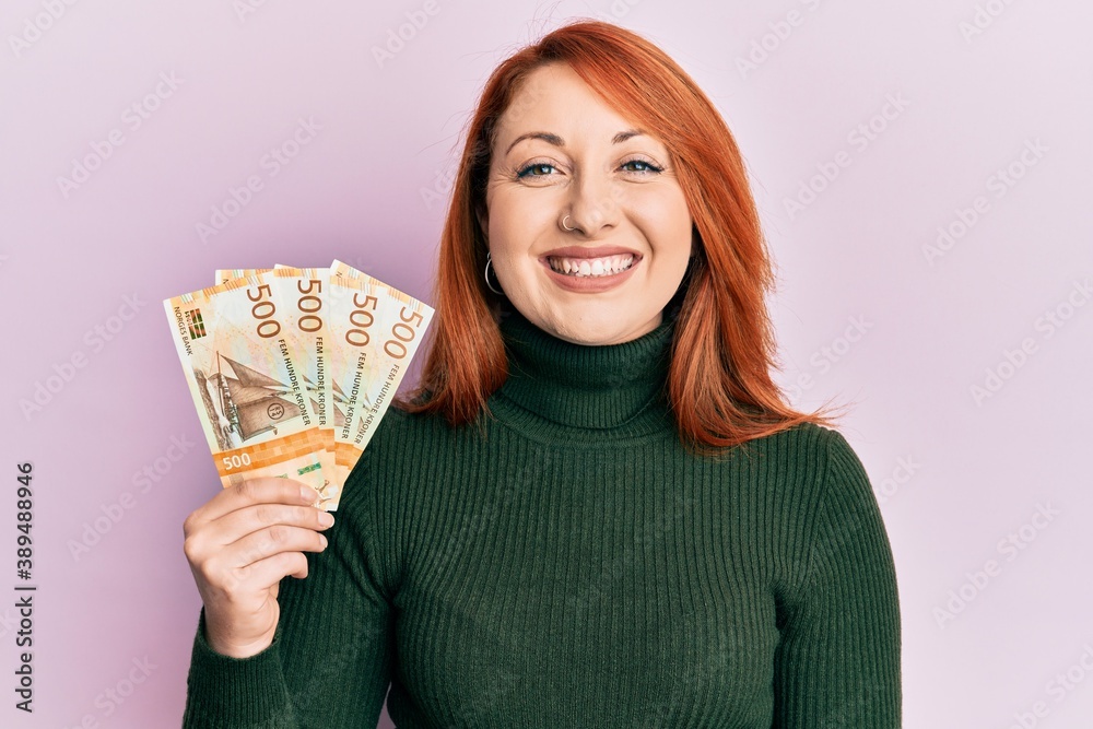 Wall mural Beautiful redhead woman holding 500 norwegian krone banknotes looking positive and happy standing and smiling with a confident smile showing teeth