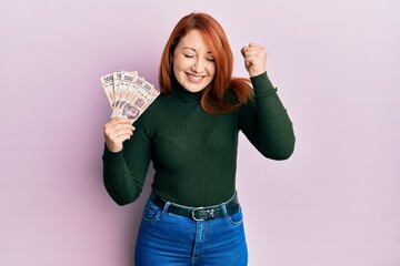 Beautiful redhead woman holding 500 mexican pesos banknotes screaming proud, celebrating victory...