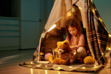 A little girl wearing pyjamas sitting on a floor cross-legged barefoot in a self-made hut made of a plaid making a massage to her fluffy friend - teddybear with fancy garlands around the hut © Svitlana