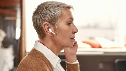 Obraz premium A sideview of a mature well-dressed woman with a short haircut and wireless earphones talking on a phone