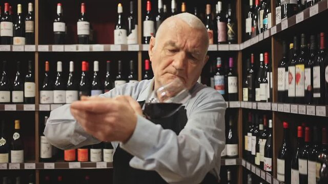 Confident elderly male winemaker holding glass of red wine, checking it in wine store