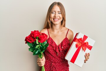 Beautiful young blonde woman holding anniversary present and bouquet of flowers smiling with a happy and cool smile on face. showing teeth.