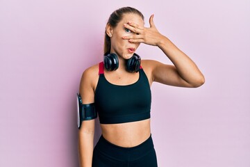 Beautiful young blonde woman wearing gym clothes and using headphones peeking in shock covering face and eyes with hand, looking through fingers afraid