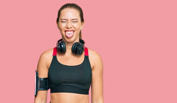 Beautiful blonde woman wearing gym clothes and using headphones sticking tongue out happy with funny expression. emotion concept.