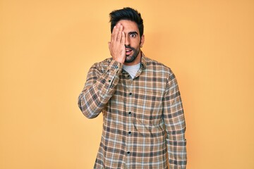 Handsome hispanic man with beard wearing casual clothes covering one eye with hand, confident smile on face and surprise emotion.