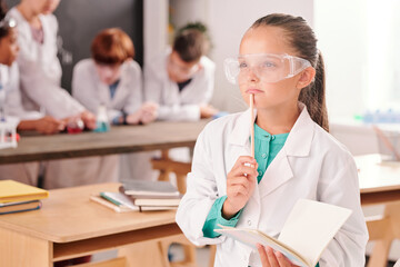 Pensive cute schoolgirl in whitecoat and protective eyeglasses holding copybook