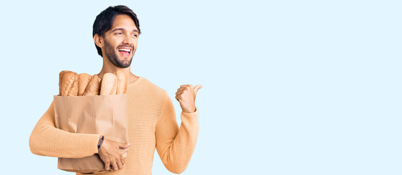 Handsome hispanic man holding paper bag with bread pointing thumb up to the side smiling happy with open mouth