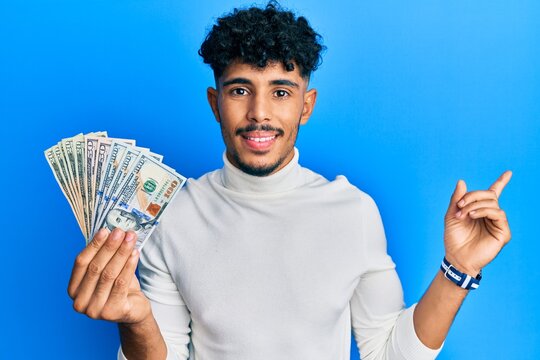 Young arab handsome man holding dollars smiling happy pointing with hand and finger to the side