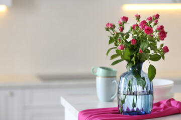 Glass vase with fresh flowers and towel on table in kitchen. Space for text